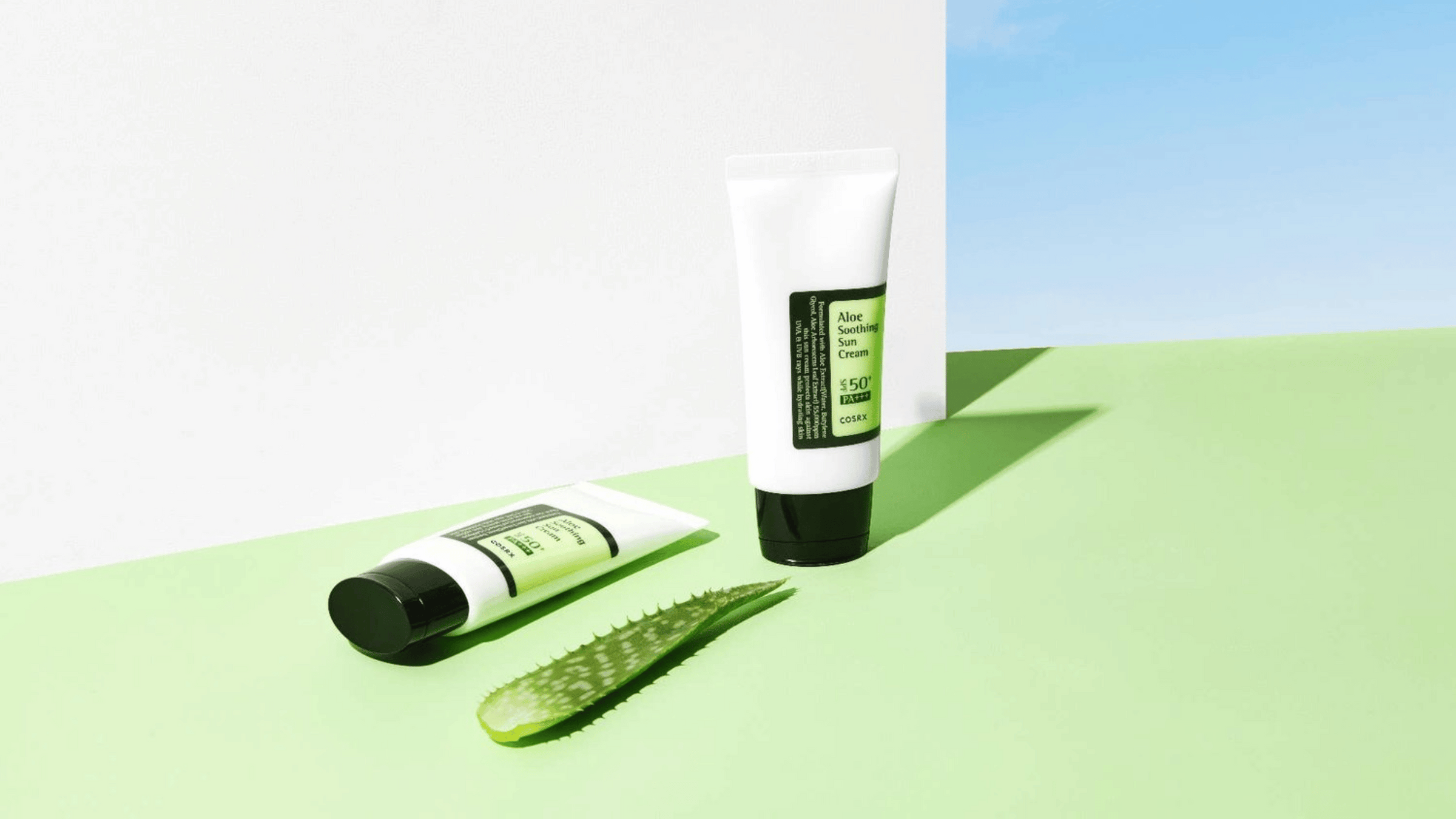 Två tuber med COSRX Aloe Soothing Sun Cream SPF50+ PA+++ står mot en ljusgrön bakgrund tillsammans med en bit färsk aloe vera och blå himmel i bakgrunden.
