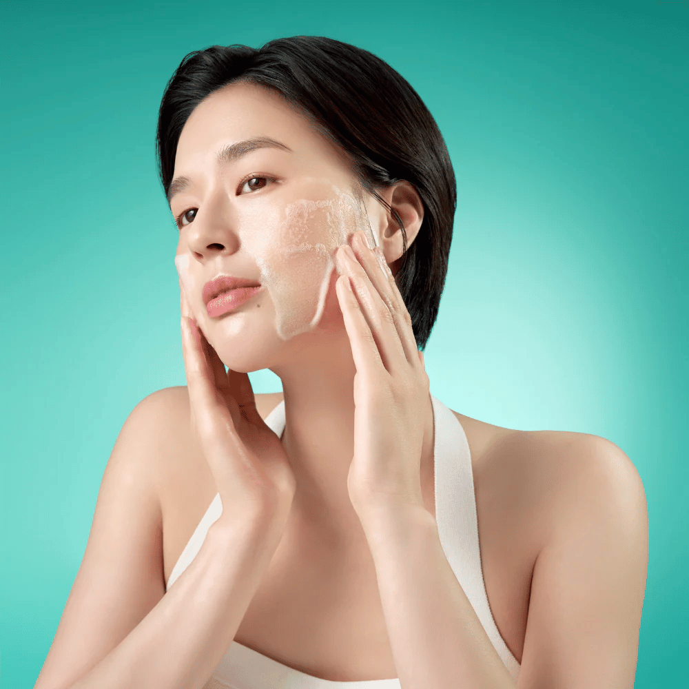 Kvinna rengör ansiktet med ett milt löddrande ansiktsrengöringsskum mot en grön bakgrund.