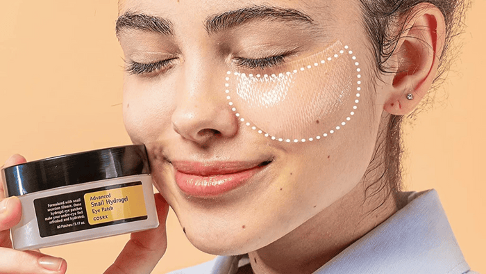 Kvinna som håller i en burk med eye patches från COSRX. Hon har även applicerat eye patches under högra ögat och ser ut att njuta av behandlingen.