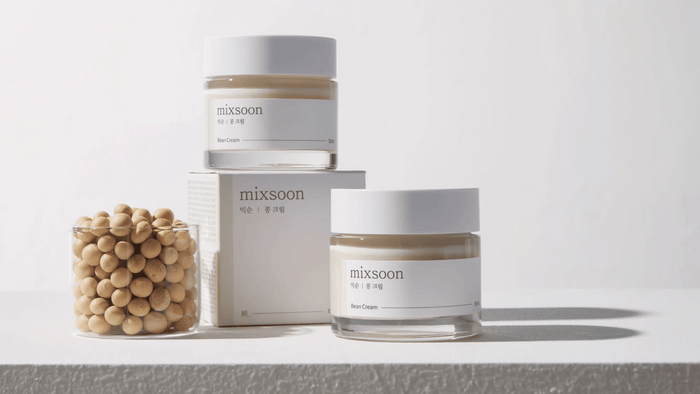 Två burkar av Mixsoon Bean Cream står på en ljus yta med en glasbehållare fylld med beige bönor och en produktkartong bredvid. Enkel och ren design med tydliga etiketter. Bakgrunden är neutral för att fokusera på produkterna.
