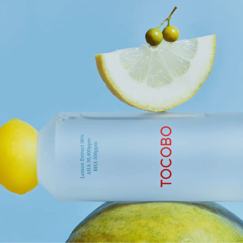 Bottle with TOCOBO AHA BHA Lemon Toner, decorated with a lemon-shaped cap. A lemon slice and small green berries balance on top of the bottle against a blue background.