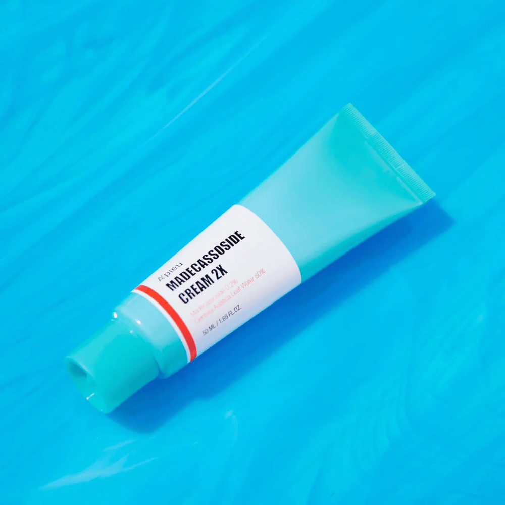 A tube A'PIEU Madecassoside Cream 2X lies diagonally over a blue background with water-like wrinkles. The tube has a white body with a light turquoise cap and a red to orange colored label where the product name and the active ingredient Madecassoside 2% are clearly printed. The label also states the quantity which is 50 ml / 1.8 fl oz. The bright and colorful presentation gives a fresh and energetic feel.