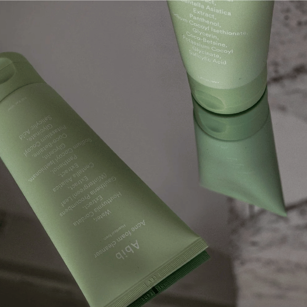 Green tube of Abib Acne Foam Cleanser Heartleaf lying on a mirrored table with the product's front and text reflected. Ingredients like salicylic acid, centella, and heartleaf extract are clearly visible on the packaging.