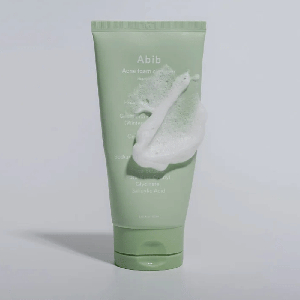 Green tube with Abib Acne Foam Cleanser Heartleaf, covered with a white foam illustrating the foaming cleansing effect. The product contains heartleaf extract and salicylic acid for acne-prone skin.
