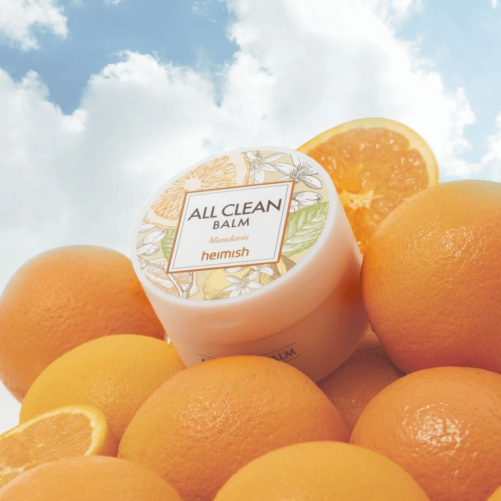 Burk av Heimish All Clean Balm Mandarin placerad på en hög med apelsiner, med en blå himmel i bakgrunden.