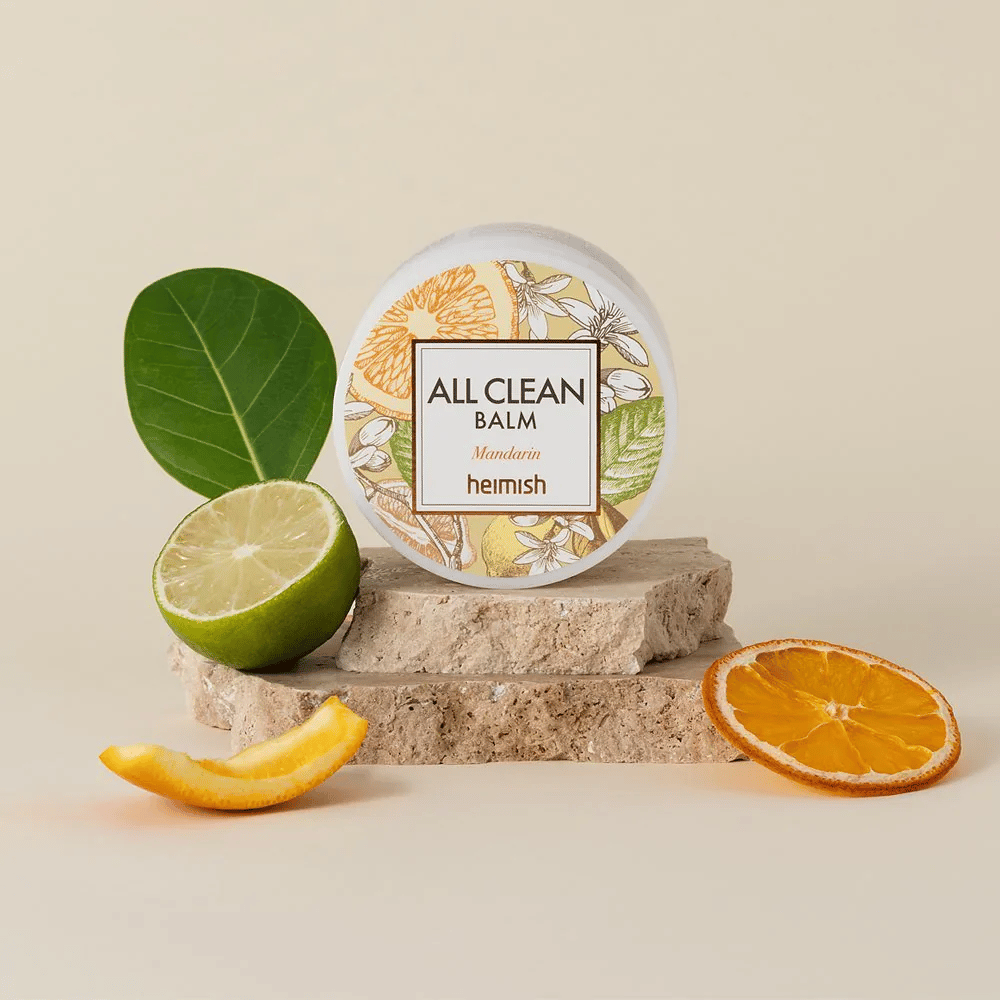 Burk av Heimish All Clean Balm Mandarin placerad på stenar, omgiven av mandarinskal och citrusblad, framför en ljus bakgrund.