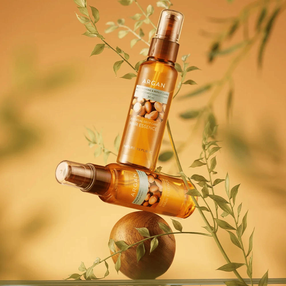 Två flaskor Nature Republic Argan Essential Moisture Hair Oil placerade bland gröna blad – silkeslen glans för torrt hår.