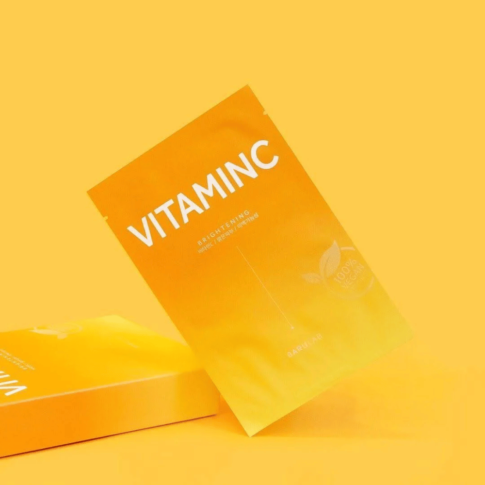 A pile of packages of BARULAB "VITAMINC Brightening" masks, all labeled as "100% Vegan Product". The packages have a color tone in various shades of orange and yellow, placed against a uniform yellow background.