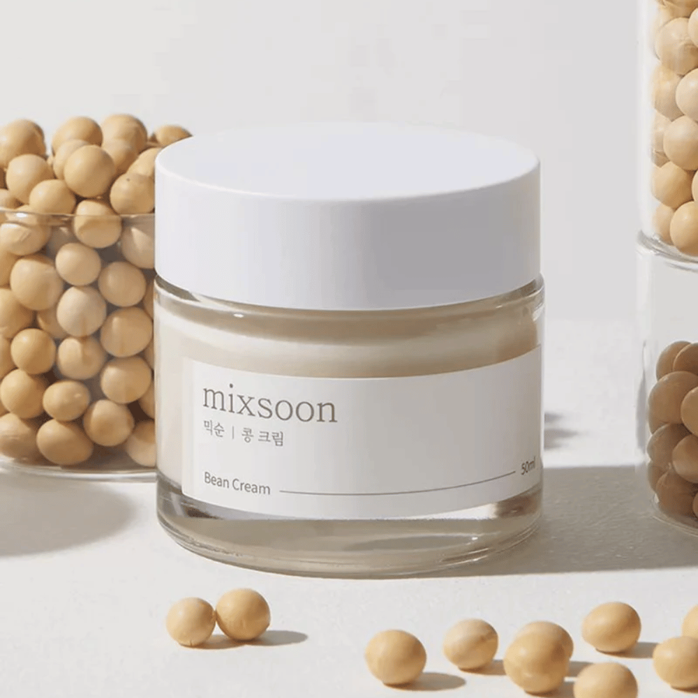 A transparent jar with a white label and lid, labeled "mixsoon 팥 크림" and "Bean Cream" with a specified volume of "50ml". The jar contains a light yellow cream and is placed on a light surface. Next to the jar is a glass container full of light brown beans that have spilled onto the surface, suggesting that the cream contains bean extract. The background and lighting provide a warm and natural atmosphere.
