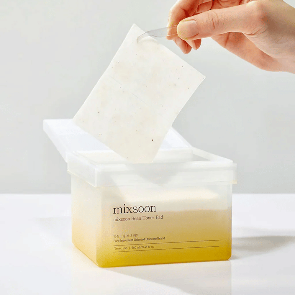 A hand holds a tweezer and lifts a Mixsoon Bean Toner Pad from a square, transparent container with a yellowish tint. The container contains 70 toner pads and has text describing the product.