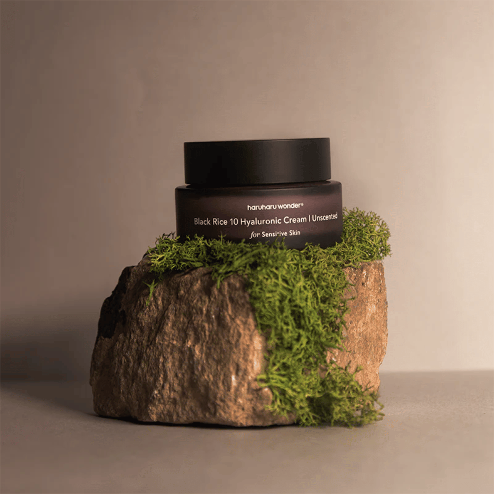 The image shows a jar of skin cream labeled "haruharu wonder Black Rice 10 Hyaluronic Cream Unscented for Sensitive Skin". The jar is black and stands on a large stone covered with light green moss, creating a natural and earthy feel.