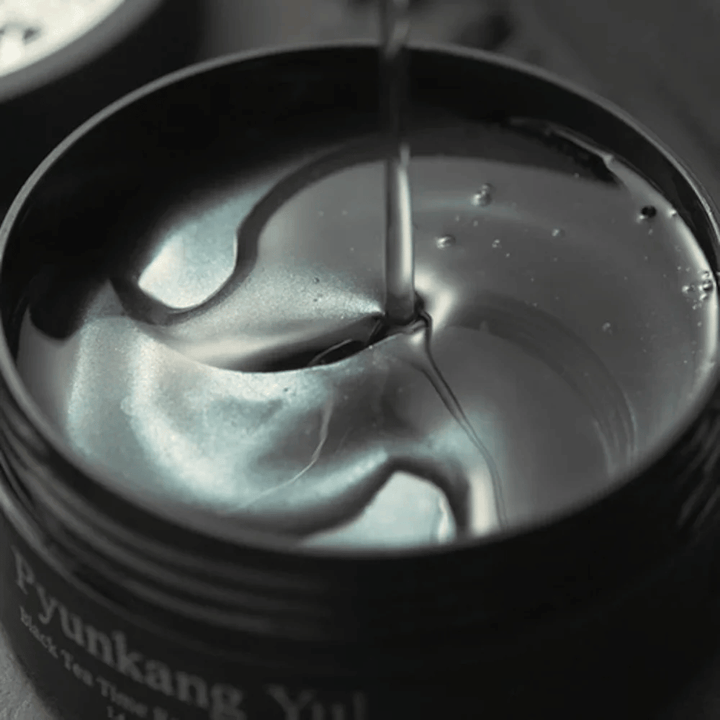 Close-up of black hydrogel eye patches in an open jar from Pyunkang Yul. The glossy patches rest in serum and highlight the product’s moisturizing and luxurious feel.