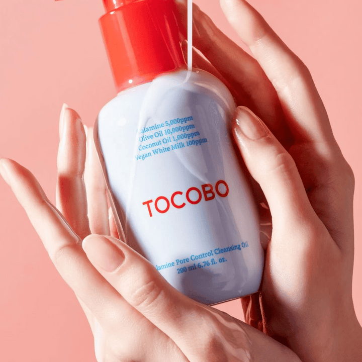 TOCOBO Calamine Pore Control Cleansing Oil hålls i händer mot en rosa bakgrund, flaskans röda pump och ingredienslista syns tydligt.
