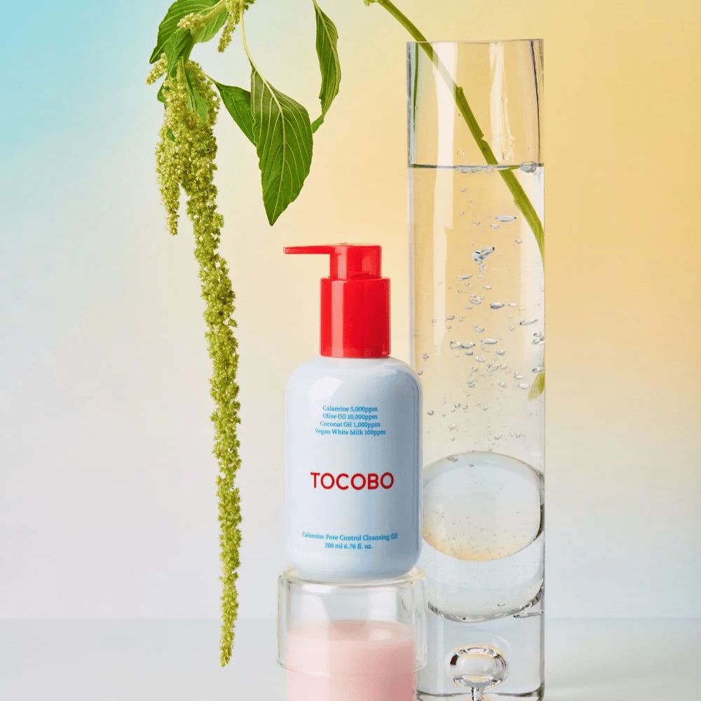 TOCOBO Calamine Pore Control Cleansing Oil med röd pump står bredvid ett högt glasrör fyllt med vatten och en grön växt, mot en ljus pastellfärgad bakgrund.