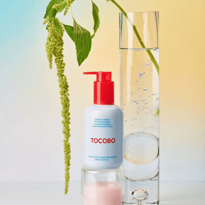 TOCOBO Calamine Pore Control Cleansing Oil med röd pump står bredvid ett högt glasrör fyllt med vatten och en grön växt, mot en ljus pastellfärgad bakgrund.