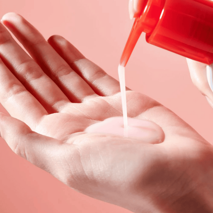En ljusrosa rengöringsolja appliceras från en röd pumpflaska ner i en öppen hand mot en rosa bakgrund.