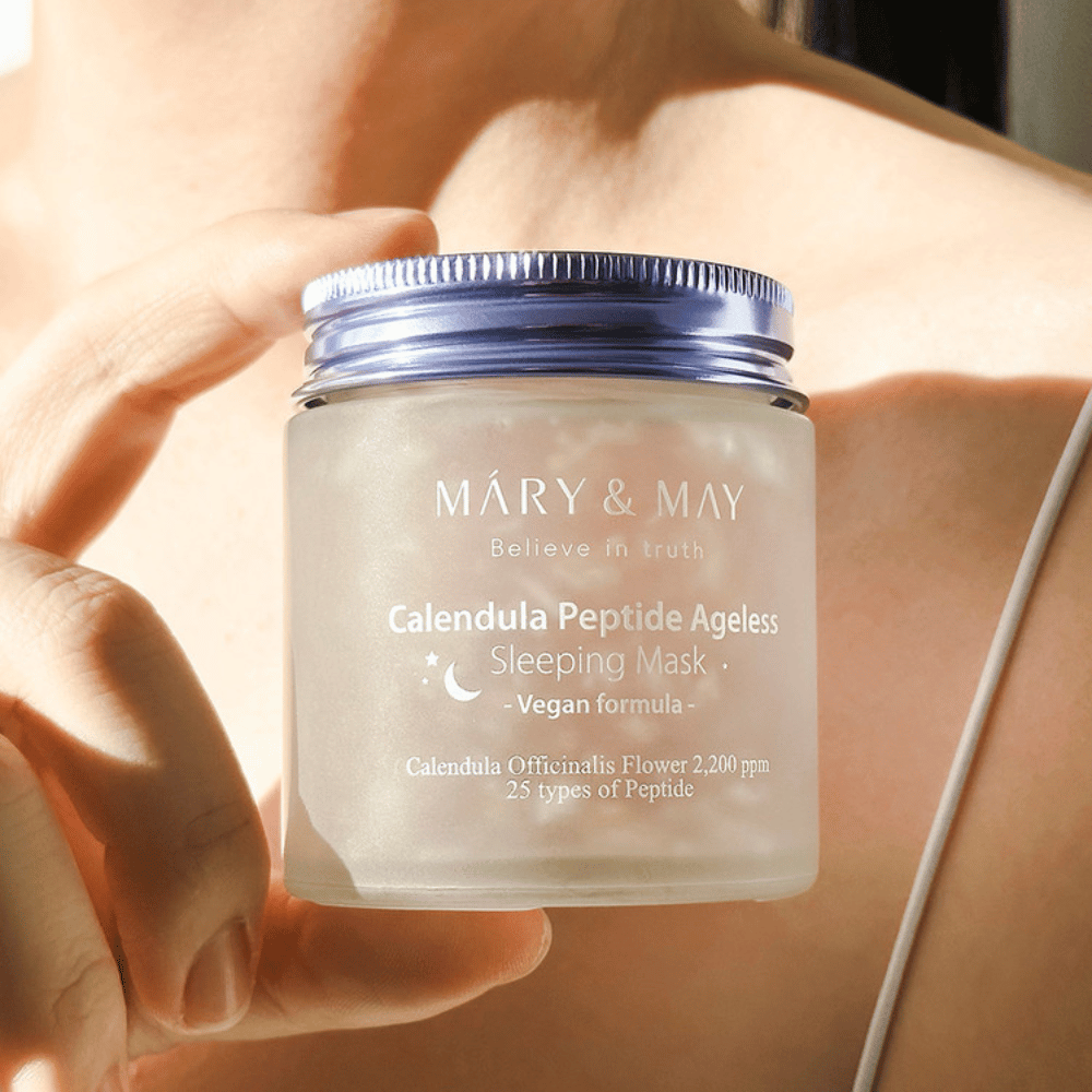 A woman holds a jar of MARY & MAY Calendula Peptide Ageless Sleeping Mask. The jar is transparent with a purple lid and the text "Calendula Peptide Ageless Sleeping Mask" is clearly visible. The product is a vegan sleeping mask with calendula and peptides, intended to give the skin a youthful glow overnight.
