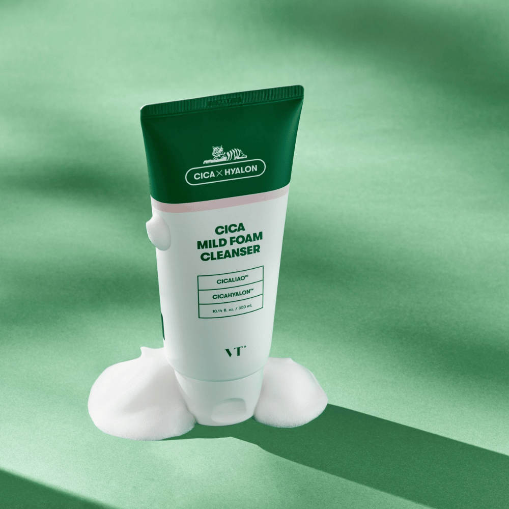 Tube with VT Cica Mild Foam Cleanser standing on a green surface with foam around the base. The product has a green and white design and is marketed with CICALIAO™ and CICAHYALON™.