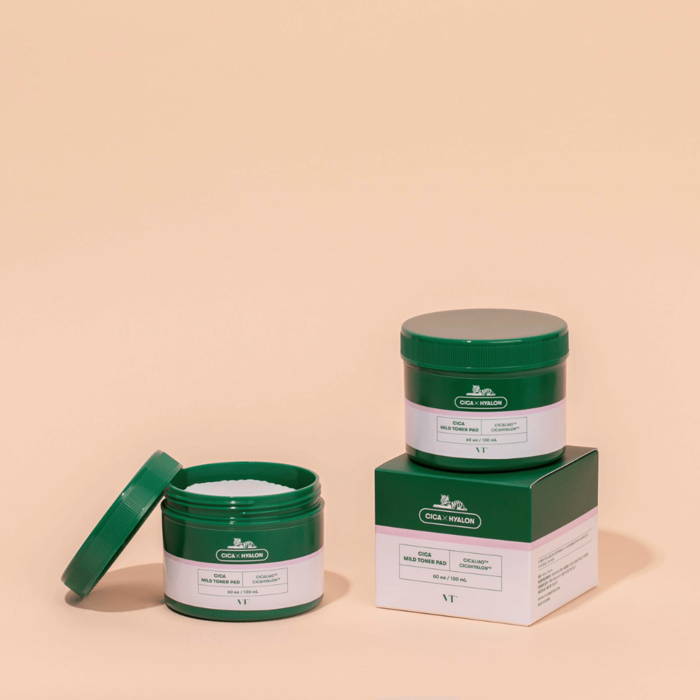 Two green jars with VT Cica Mild Toner Pad, one open showing the white toner pads and one closed placed on top of its product packaging, against a beige background.
