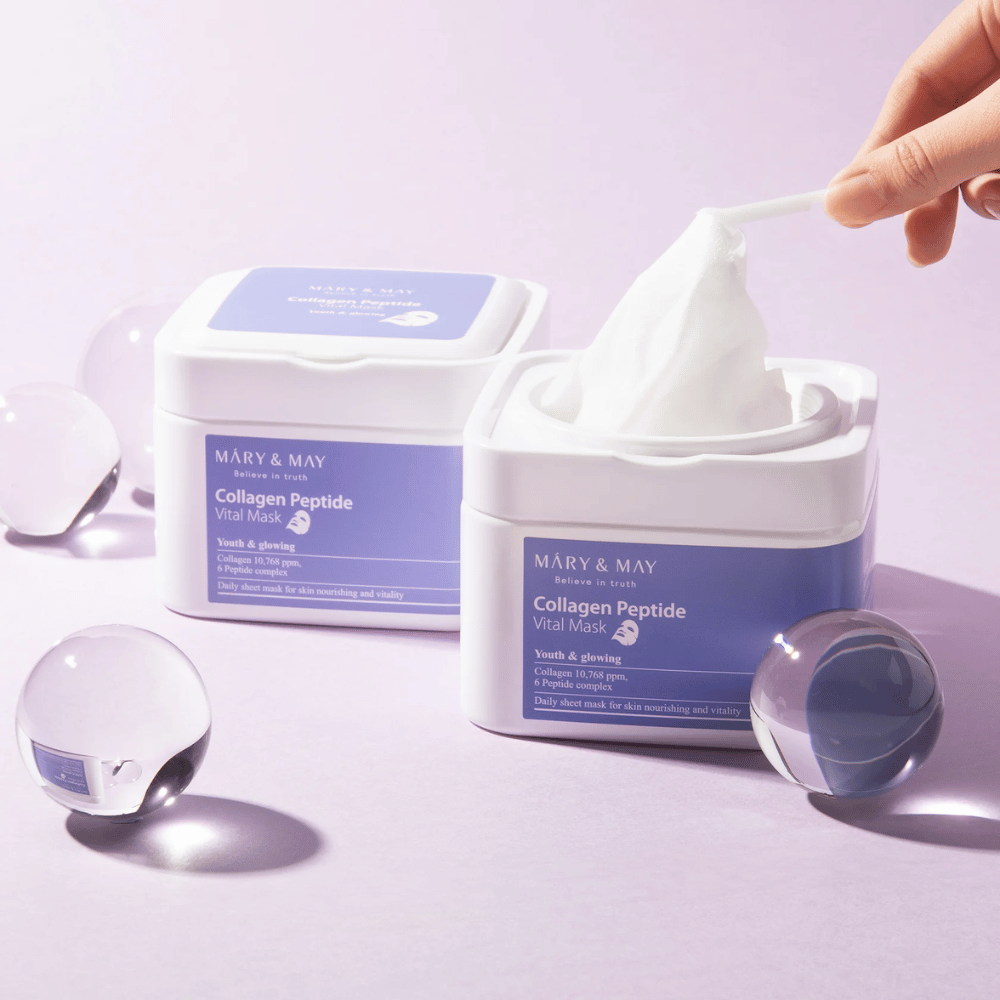 A hand picks up a mask from a jar with MARY & MAY Collagen Peptide Vital Mask. The mask is intended for daily use to nourish and glow the skin, with collagen and peptides to support the skin's youthfulness.