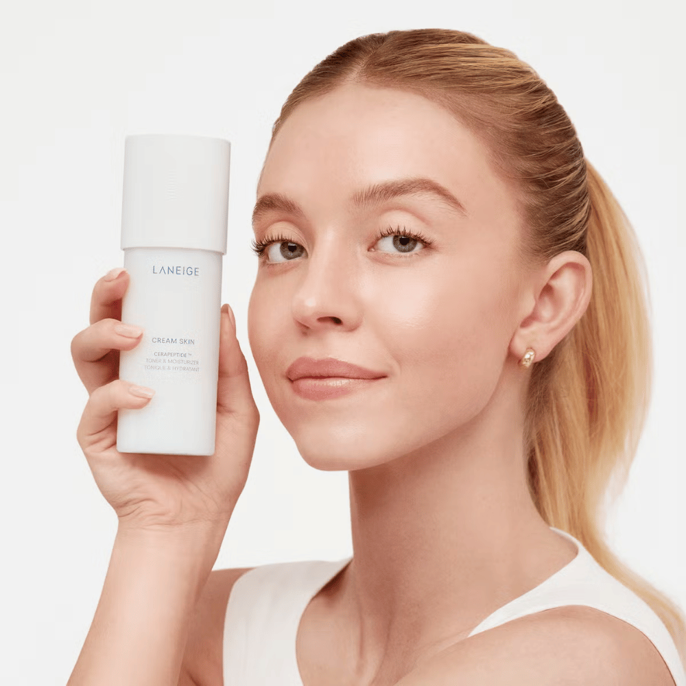 A person holds up Laneige Cream Skin Cerapeptide Refiner in a white bottle. The person smiles and shows the product, which is marketed as a moisturizing toner and skincare product.