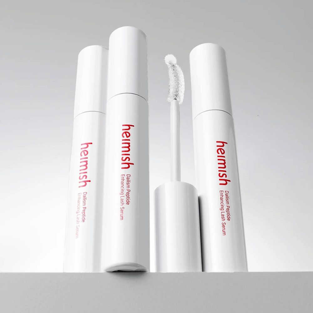 Several packages of Heimish Dailism Peptide Enhancing Lash Serum are shown together with the curved applicator brush. The image highlights the product's elegant design and precision brush for easy application.