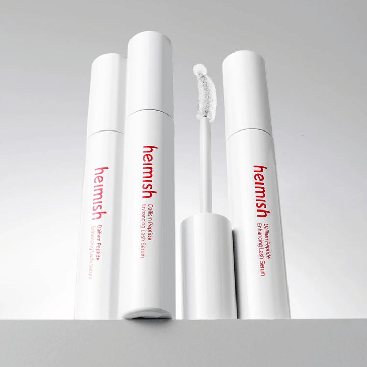 Several packages of Heimish Dailism Peptide Enhancing Lash Serum are shown together with the curved applicator brush. The image highlights the product's elegant design and precision brush for easy application.