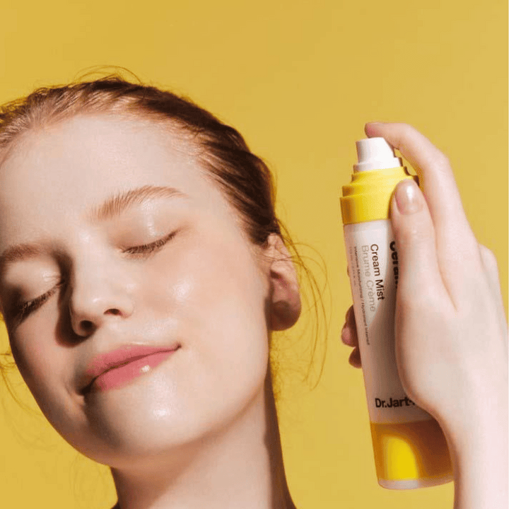 En kvinna håller en flaska Dr. Jart+ Ceramidin Cream Mist 110ml i ansiktet, vilket främjar dess fukt-boost-fördelar och vegansk formulering.