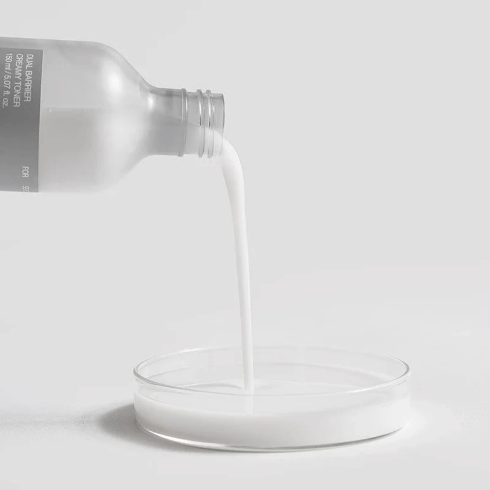 Celimax Dual Barrier Creamy Toner is gently poured from the bottle into a round laboratory well, with its milky and nutrient-rich formula clearly in focus.