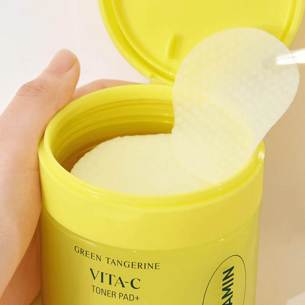 A hand uses a plastic tweezer to pick up a textured toner pad from a light yellow container labeled "GREEN TANGERINE VITA-C TONER PAD+" by the brand "gooda