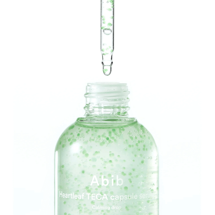 Minimalist close-up of serum bottle and dropper where the transparent formula with green capsules is the focus against a light background.