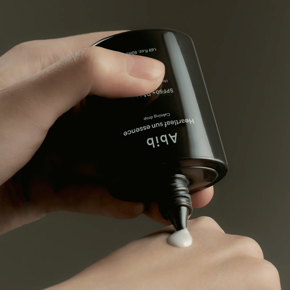 Hand squeezing out Abib Heartleaf Sun Essence Calming Drop on the back of another hand, with a dark background and a clear display of the product's texture.