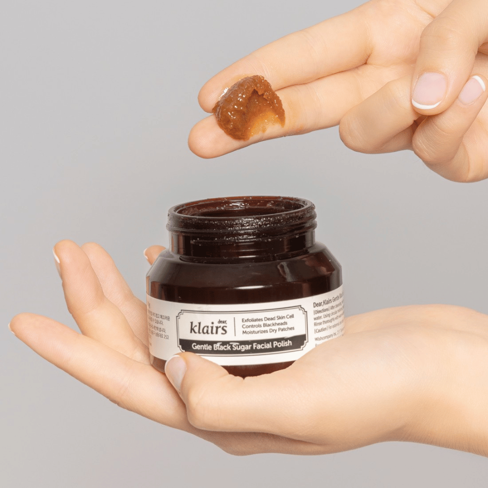 Hands holding an open jar of Klairs Gentle Black Sugar Facial Polish and showing the product on a finger, against a gray background.