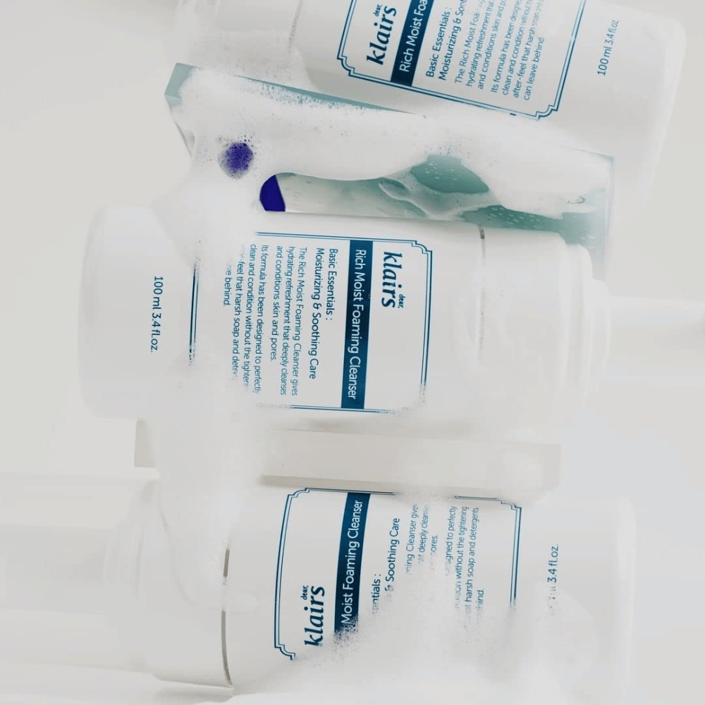 Three bottles of Klairs Rich Moist Foaming Cleanser covered with foam and water droplets, placed on a white surface.