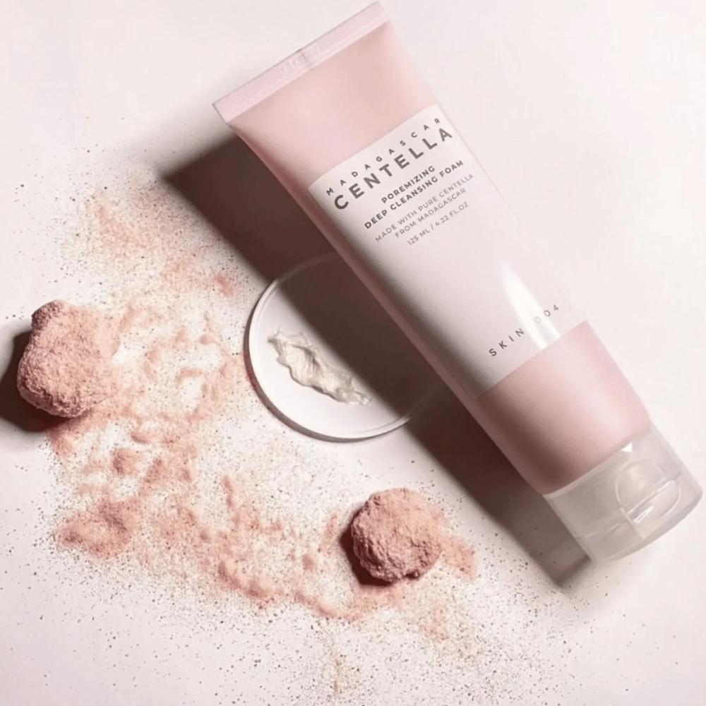 Madagascar Centella Poremizing Deep Cleansing Foam is presented in a pink tube. The tube is placed on a light surface along with pink powder that gives a sense of purity and freshness. The product is designed to deeply cleanse the skin using pure centella from Madagascar.