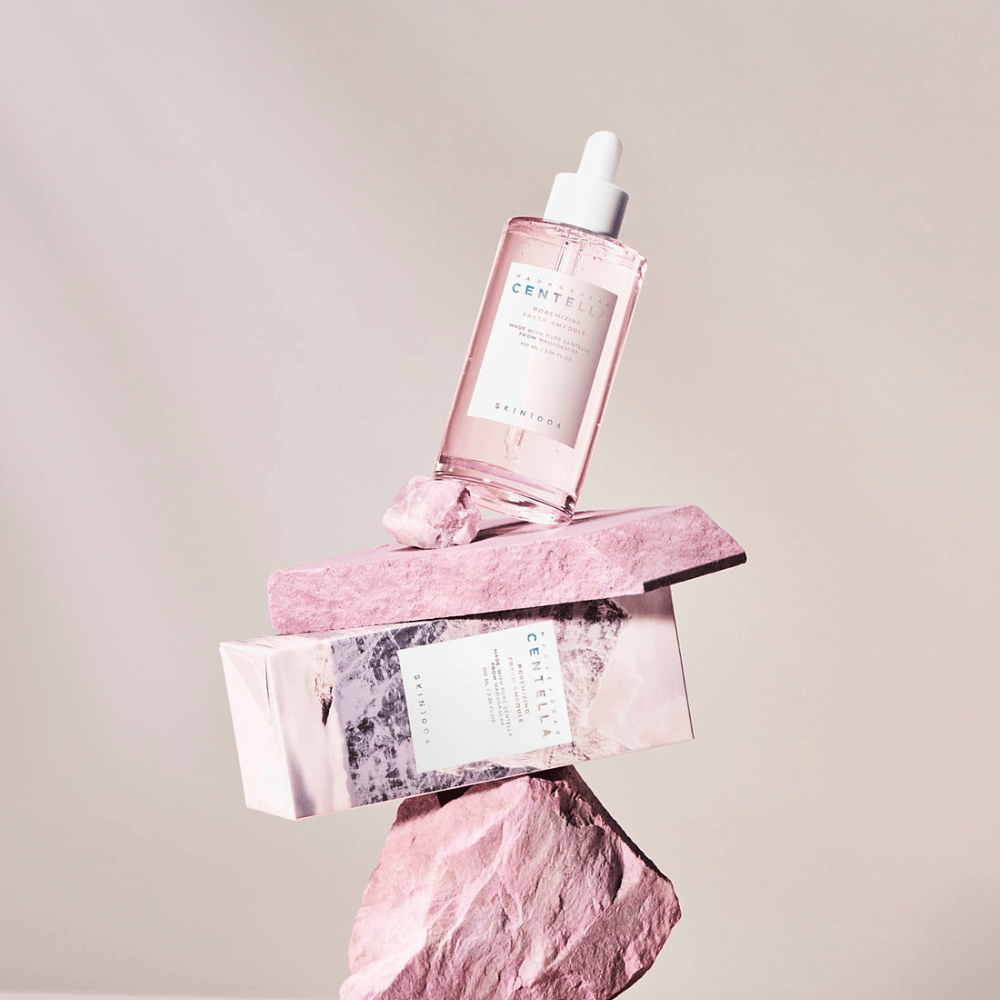 Transparent bottle with pink Centella ampoule from SKIN1004, balanced on stone blocks, with a cardboard packaging underneath, against a neutral background.