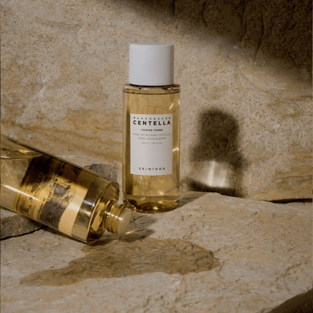 The image shows a bottle "Madagascar Centella Toning Toner" from SKIN1004. It is a transparent bottle filled with a yellowish liquid, presumably a toner for skincare. It states that it is made with pure Centella from Madagascar and the content is 210 ml / 7.10 fl.oz. The bottle and a lying bottle are placed on a rocky surface, providing a natural and minimalist feel.