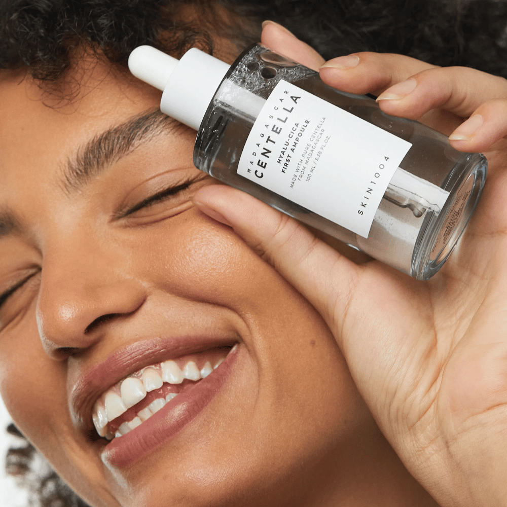 Smiling woman holds up Skin1004 Madagascar Centella Hyalu-Cica First Ampoule next to her face.