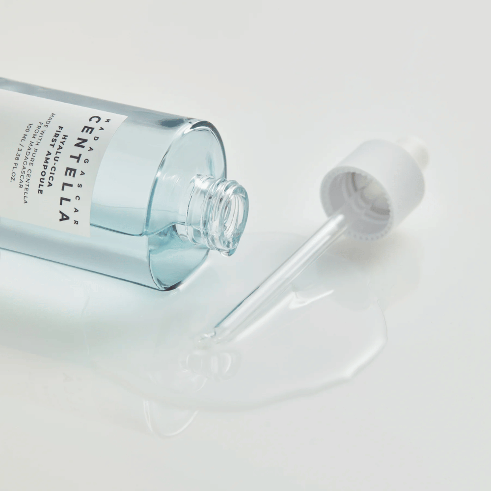 Close-up of open bottle Skin1004 Madagascar Centella Hyalu-Cica First Ampoule with dropper lying down and serum spread on white surface.