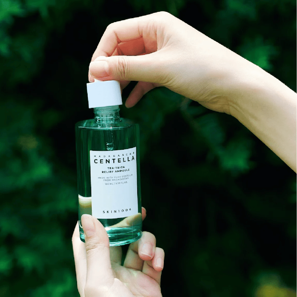 Person öppnar SKIN1004 Madagascar Centella Tea-Trica Relief Ampoule – grön glasflaska med vit etikett hålls i händer framför grönskande bakgrund.