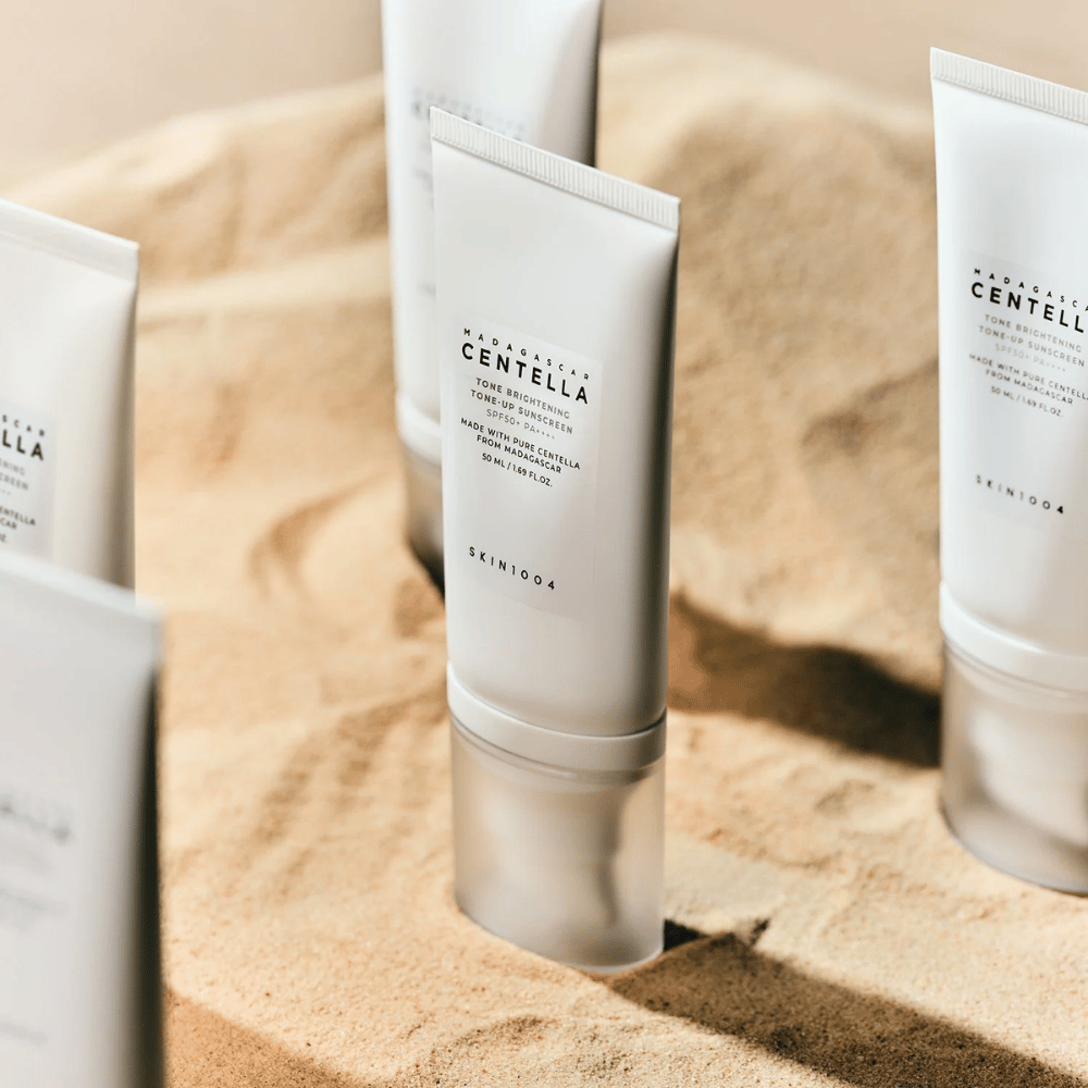 Several tubes of Madagascar Centella Tone Brightening Tone-Up Sunscreen stand in the sand. The tube is white with black text and contains 50 ml. The product has SPF 50+ PA++++ and is made with pure Centella from Madagascar.