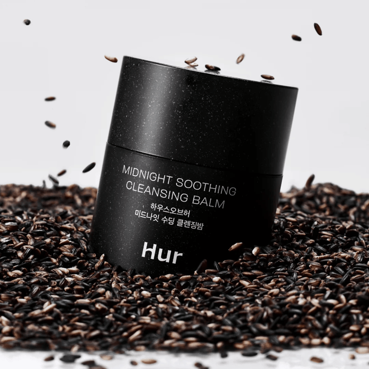 How Midnight Soothing Cleansing Balm in black jar placed on black seeds. Soothing cleansing balm that dissolves makeup and impurities while caring for the skin.