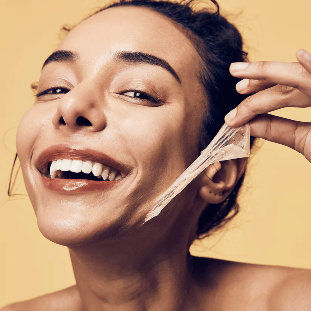 The image shows a woman with a big smile removing a peel-off mask from her face. The woman looks happy and satisfied while using the beauty product, conveying a sense of well-being and skincare.