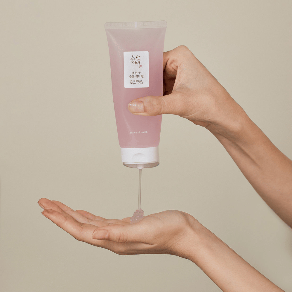 The image shows a person holding a tube of "Red Bean Water Gel" from Beauty of Joseon and squeezing out a transparent gel into their hand. The product appears to be intended for skincare and has an attractive, soft pink packaging.