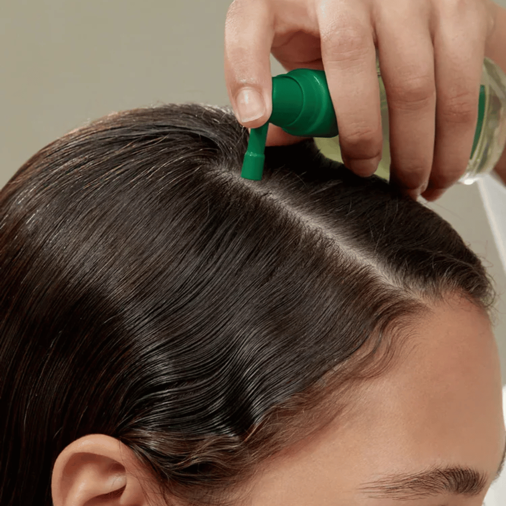 A person applies AROMATICA Rosemary Root Enhancer directly to the scalp using the targeted nozzle. The image shows how the product is used for scalp care and hair root strengthening.