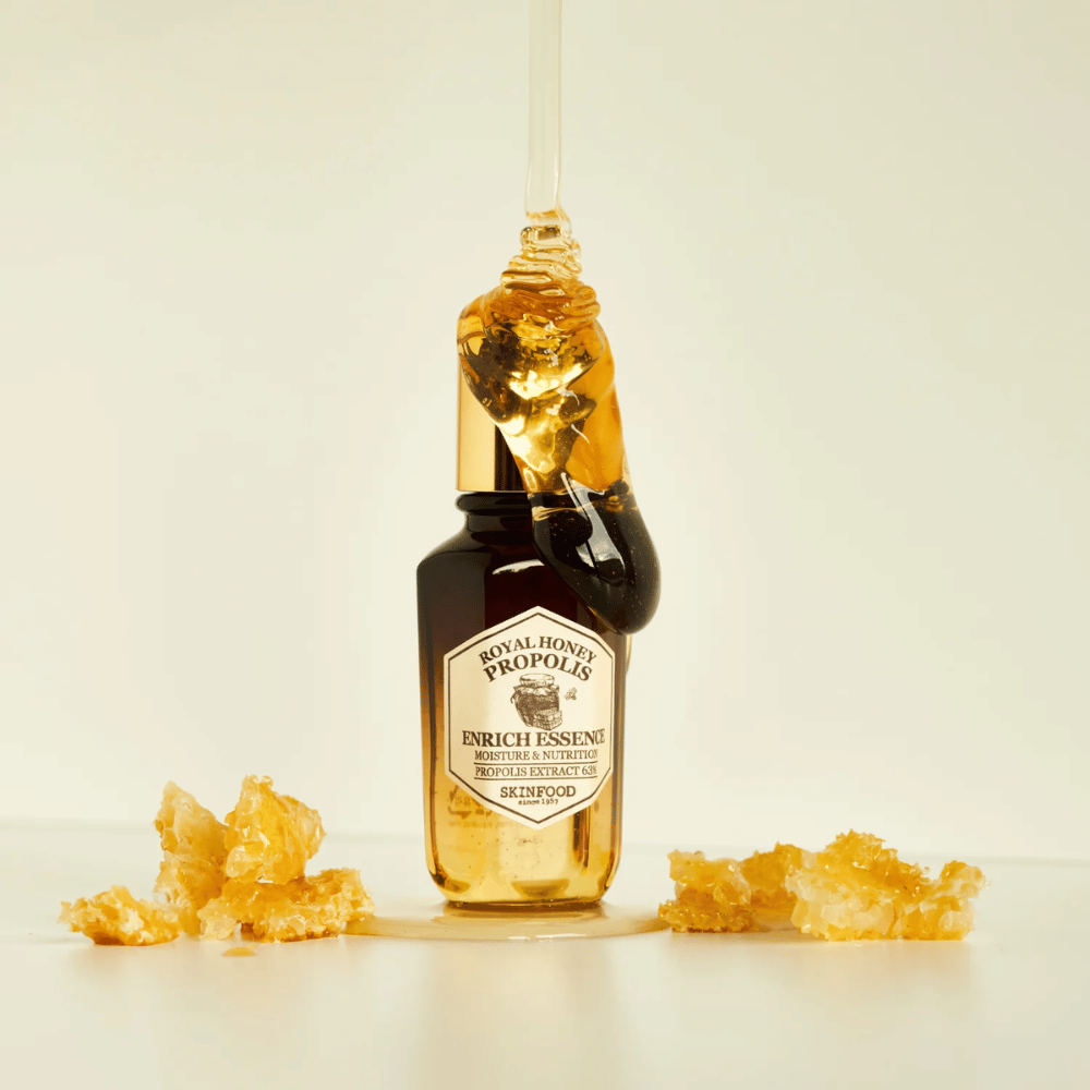 Image of a glass bottle with Royal Honey Propolis Enrich Essence from Skinfood. A large drop of golden honey runs down from the bottle's gold-colored cap. Around the bottle, small pieces of honeycomb are scattered on a light surface. The essence is marketed with a focus on moisture and nutrition, with 63% propolis extract.