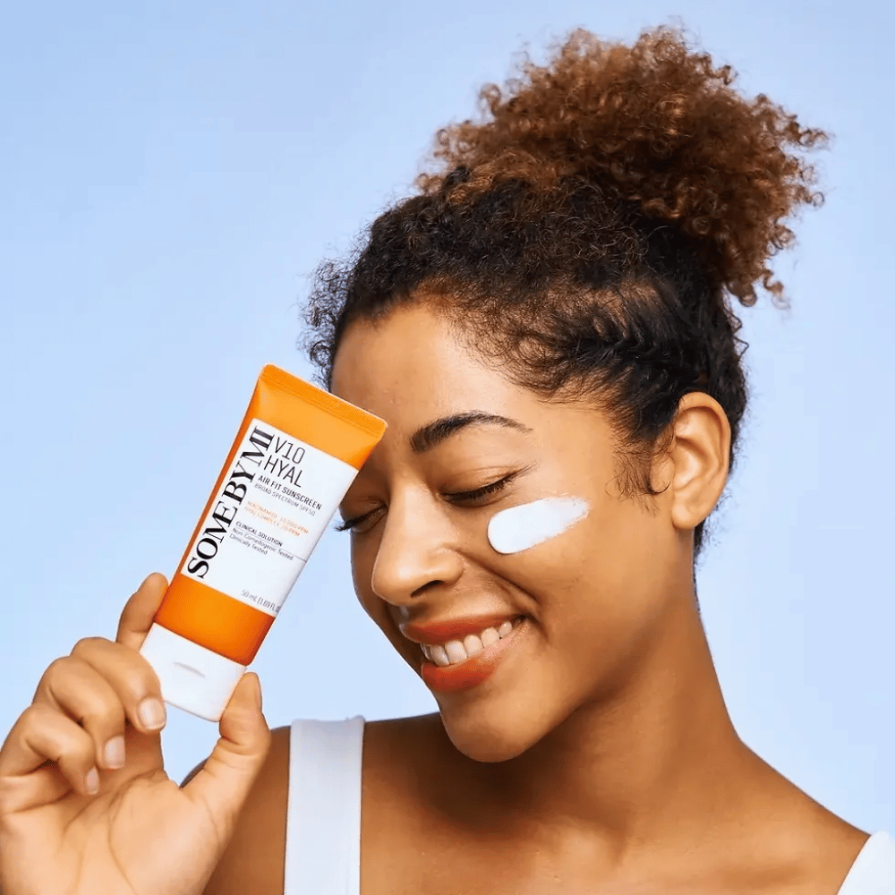 A woman holds a tube of SOME BY MI V10 Hyal Air Fit Sunscreen and has a white spot of sunscreen on her cheek. She smiles happily and clearly shows the product, highlighting its use and benefits for skincare and sun protection.