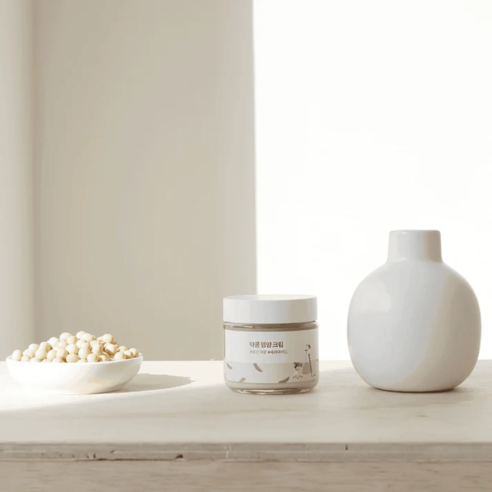 Mixsoon Soybean Nourishing Cream stands on a wooden table next to a vase and soybeans. A minimalist, moisturizing cream with natural ingredients.