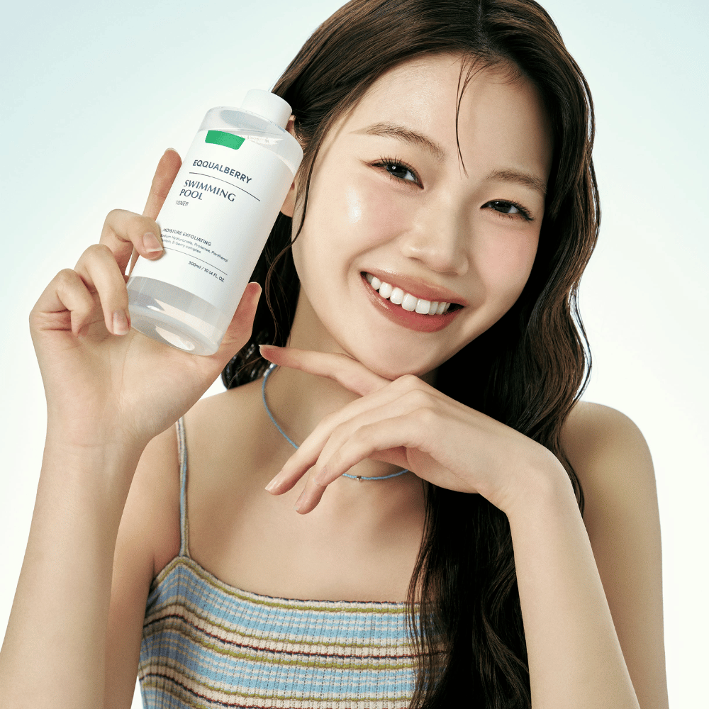 Smiling woman with radiant skin holding up Equalberry Swimming Pool Toner. Fresh and natural look with light makeup and a summery outfit in blue tones.