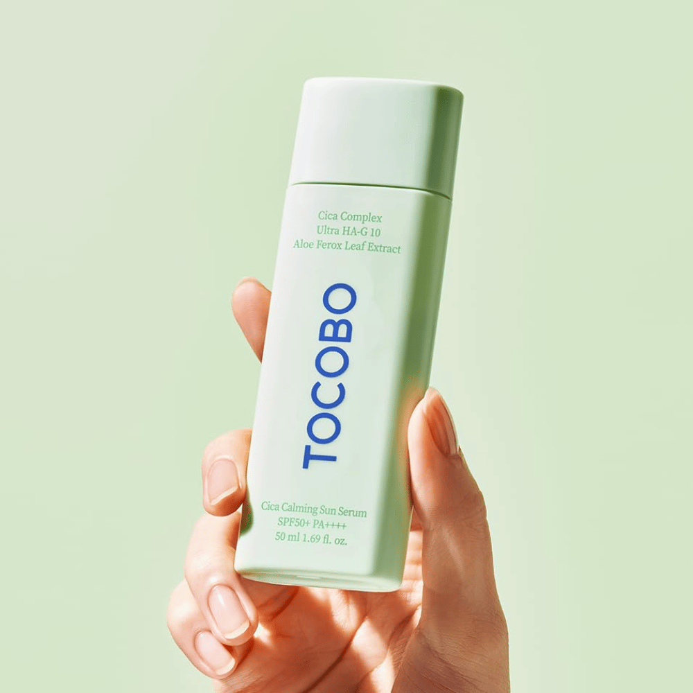 Hand holding TOCOBO Cica Calming Sun Serum with mint green packaging and blue text against a light green background.